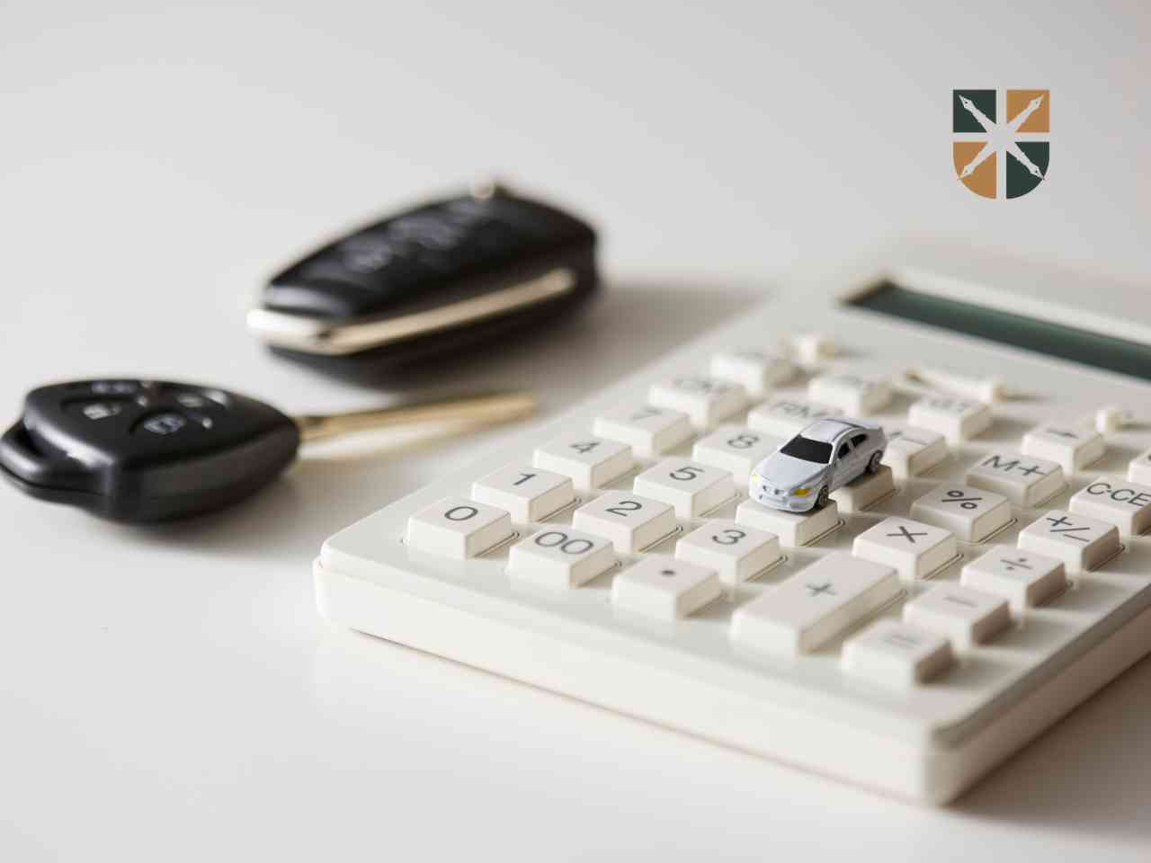 White calculator with a small silver toy car on the numeric keypad; car keys blurred in the background and a logo in the top-right corner.