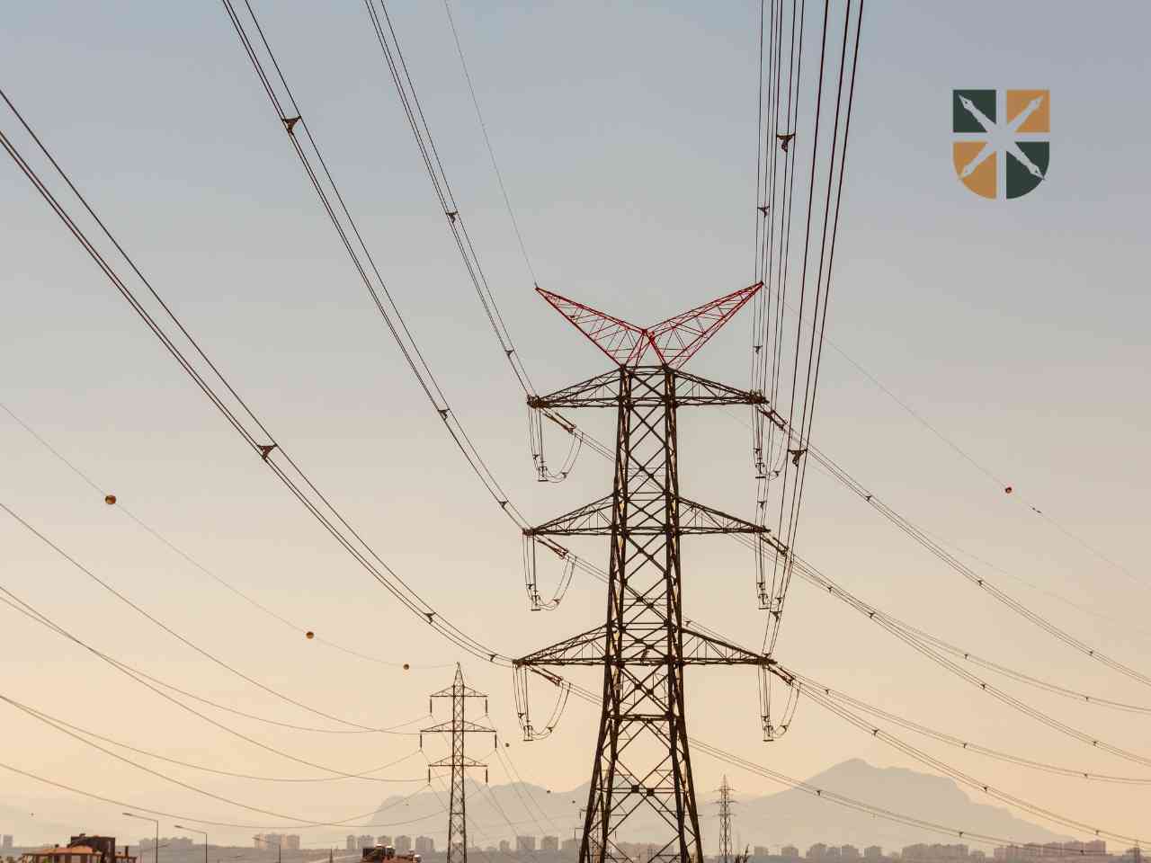 Kratowe słupy elektroenergetyczne tworzące linię wysokiego napięcia na tle zachodzącego słońca i gór.