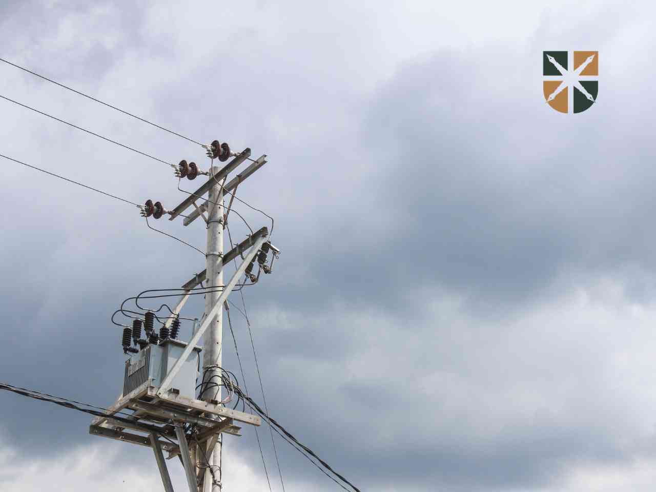 Betonowy słup energetyczny z transformatorem i liniami średniego napięcia na tle zachmurzonego nieba.