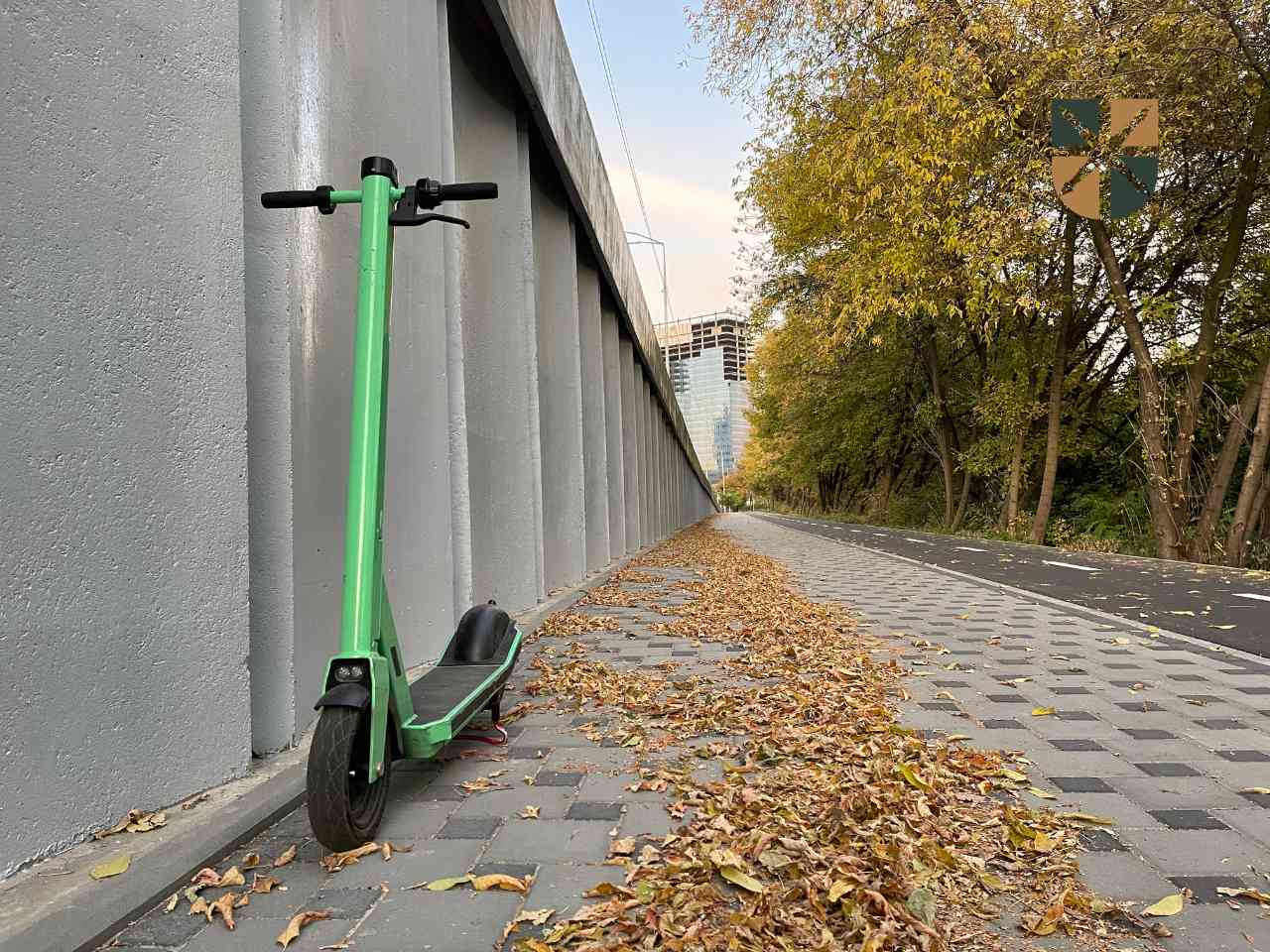 Zielona hulajnoga elektryczna zaparkowana na chodniku pokrytym jesiennymi liśćmi przy betonowej ścianie.