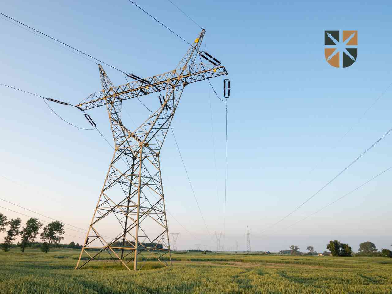 Słup energetyczny wysokiego napięcia stojący na zielonym polu pod bezchmurnym niebem.