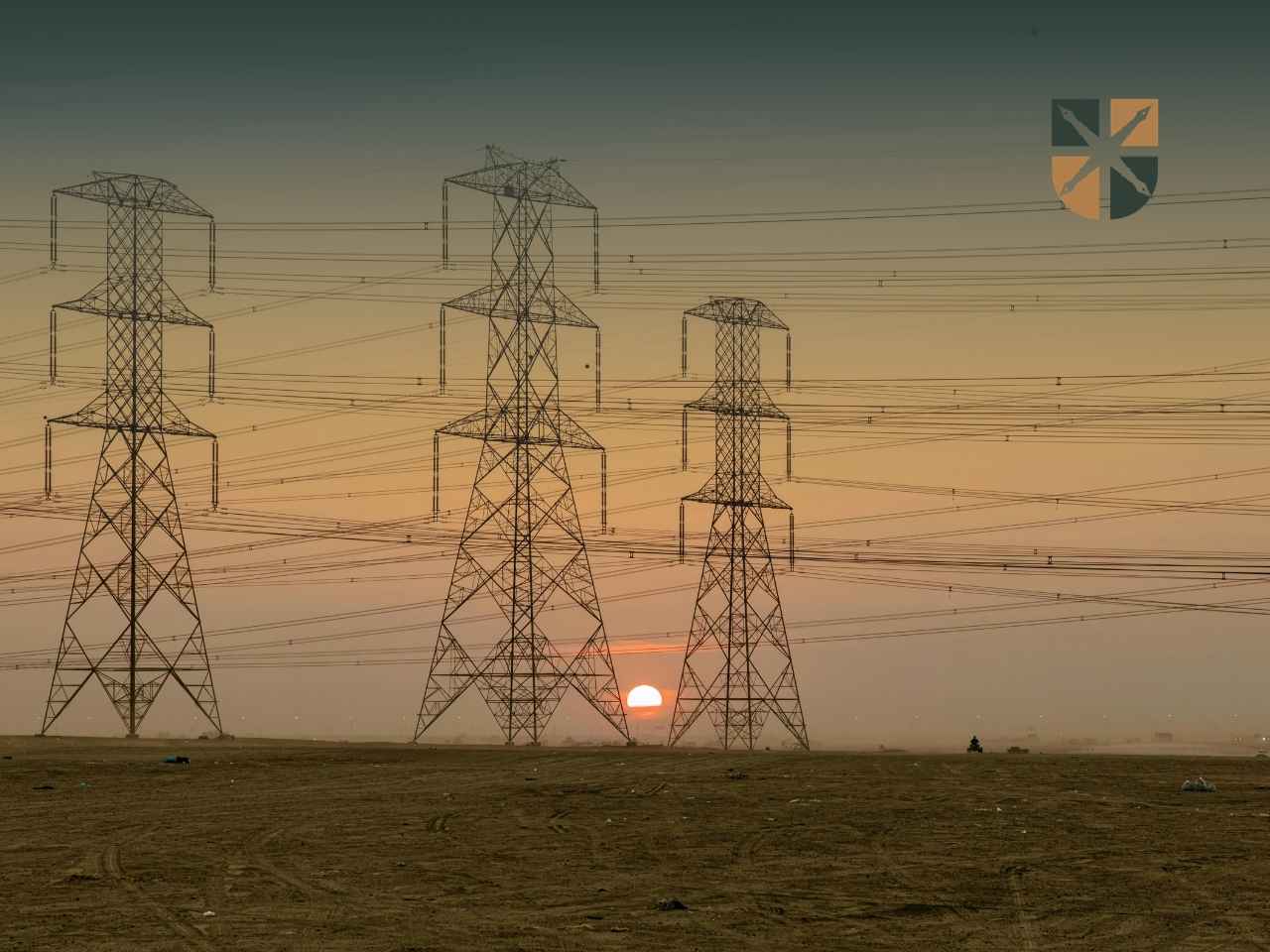 slupy-zachod-slonca Trzy duże stalowe słupy energetyczne na tle zachodzącego słońca i piaszczystego terenu.