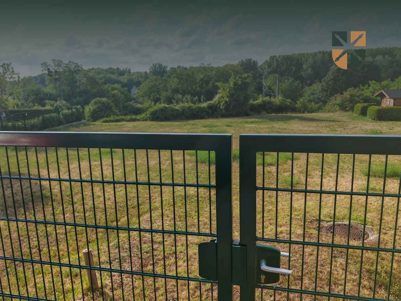 niezabudowana-dzialka Green metal gate with a latch, opening to a sunlit grassy yard and distant trees beyond the fence.